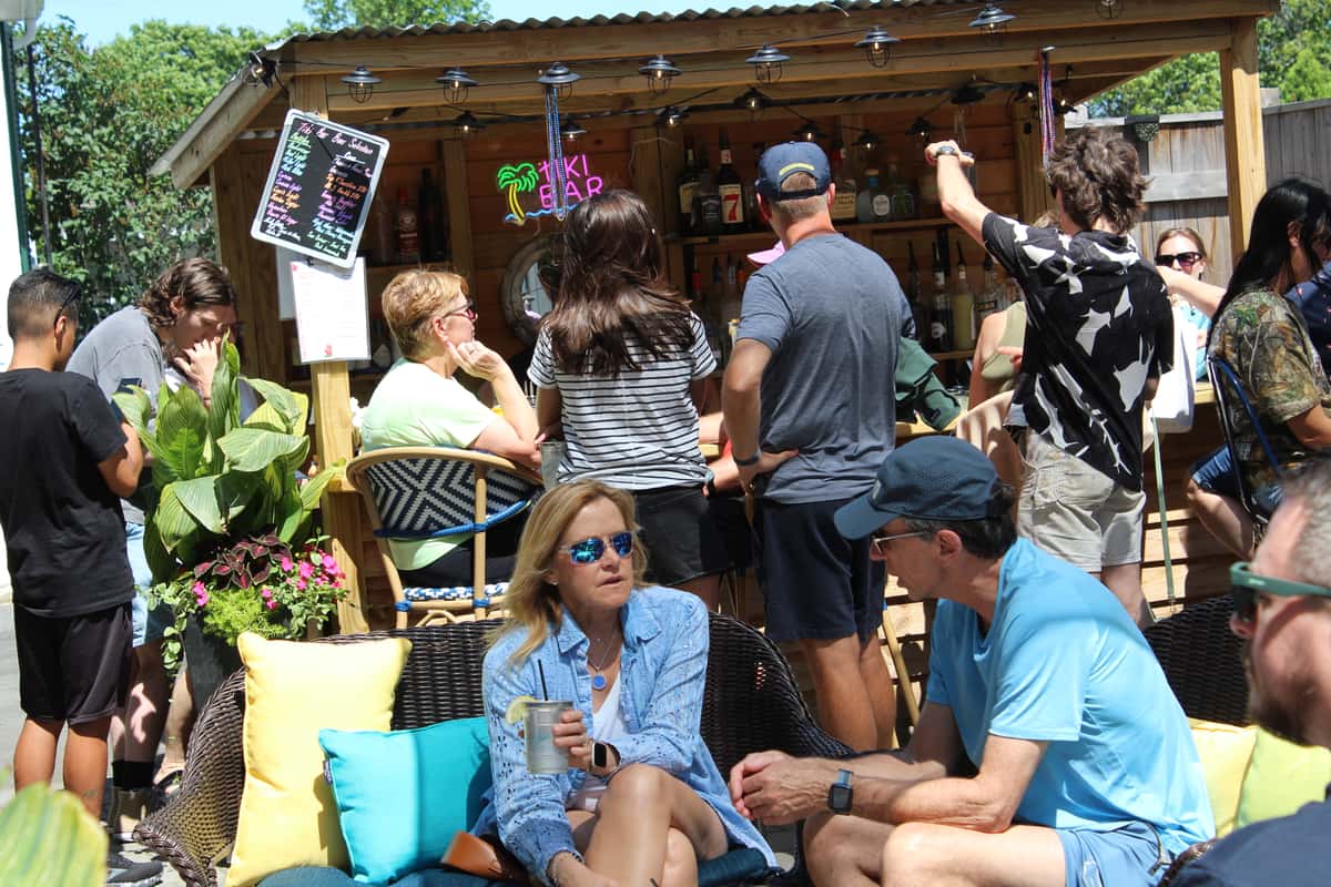 A lively outdoor tiki bar scene with people mingling, ordering drinks, and relaxing on wicker furniture with colorful pillows under the sun.