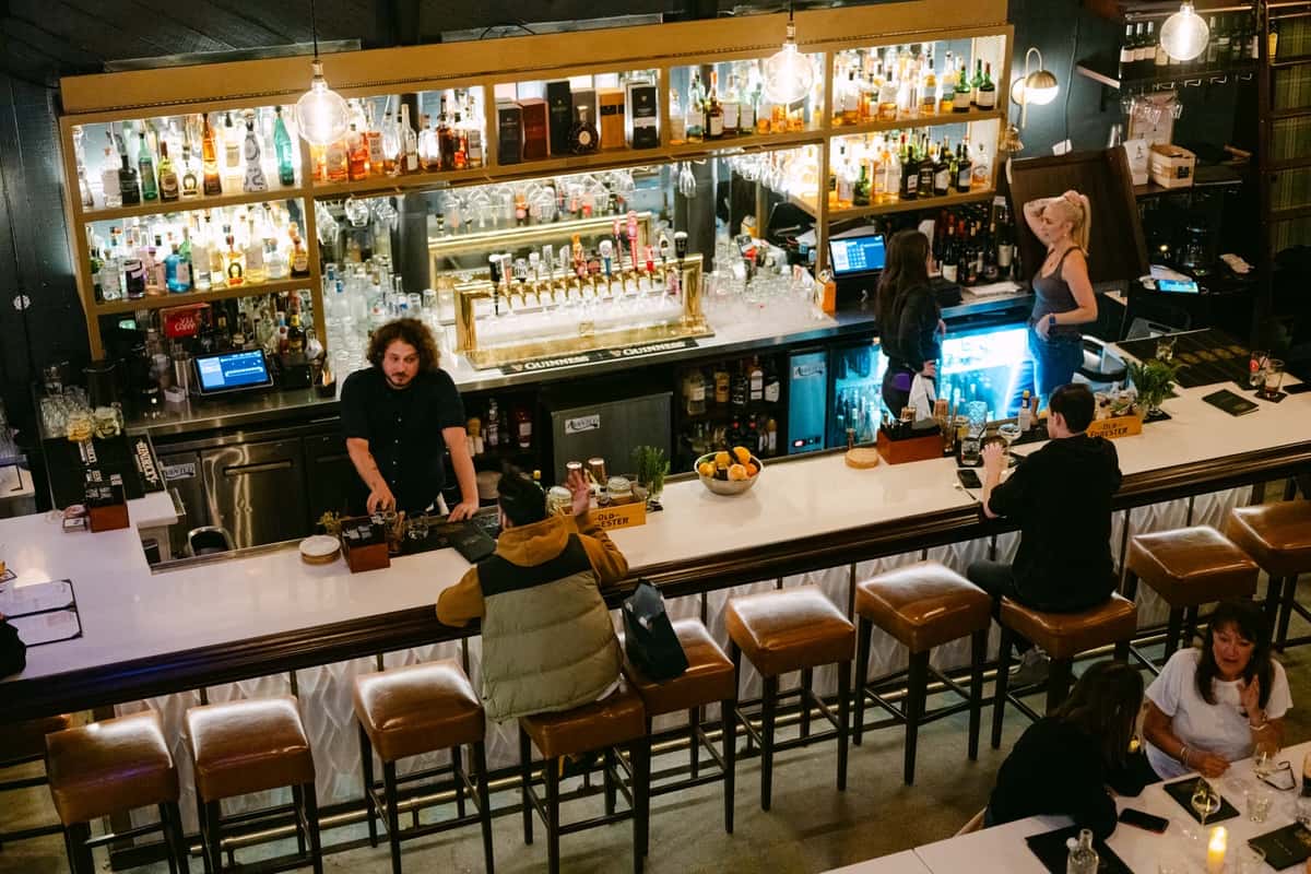 A lively bar scene with a bartender and patrons. The well-stocked bar features colorful bottles and glasses, creating an inviting atmosphere for social gatherings.