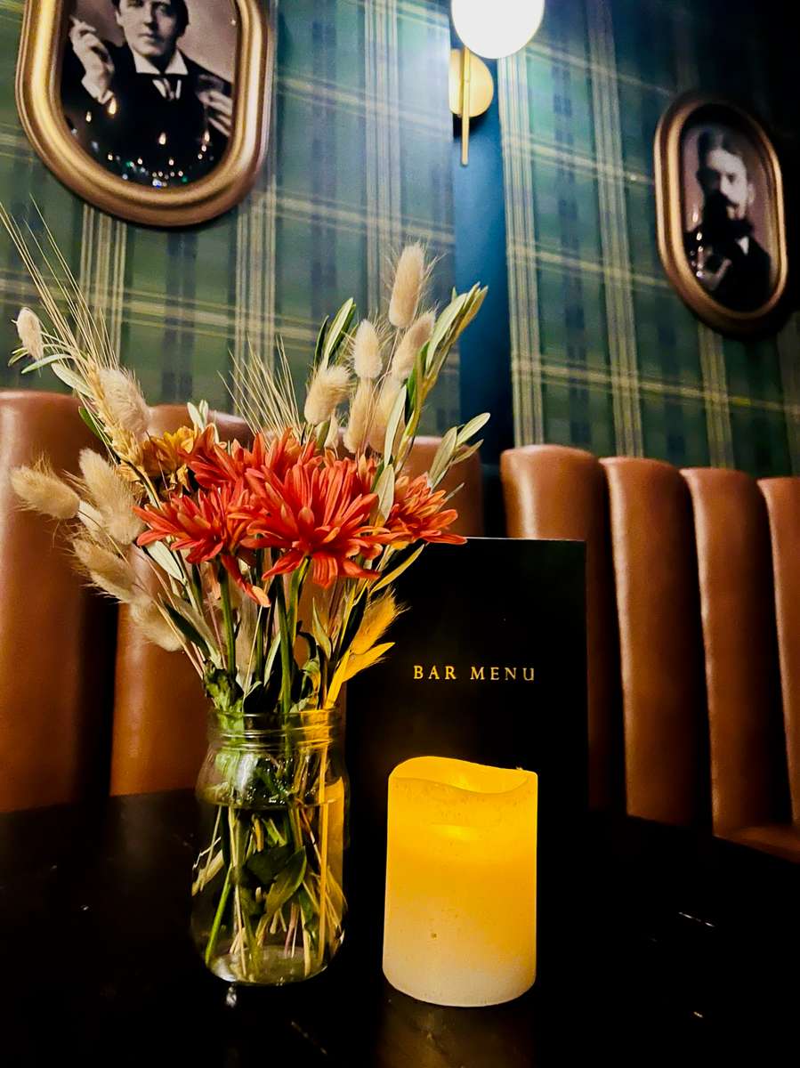 A cozy bar setting featuring a vase of colorful flowers beside a candle and a bar menu. The background has framed portraits and a patterned wall, creating a warm atmosphere.