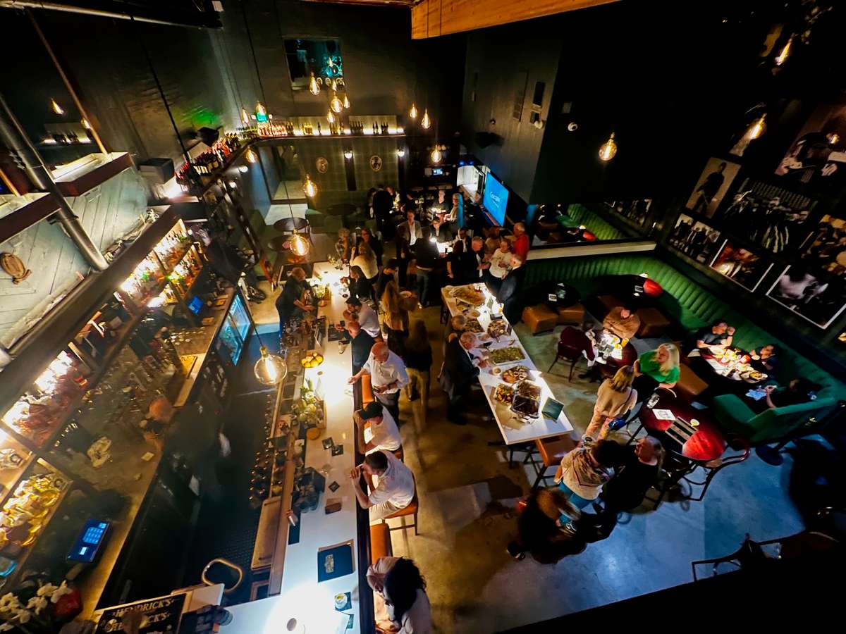 Vibrant LA bar scene with patrons enjoying drinks and food, illuminated by warm pendant lights. A lively atmosphere with a mix of seating and a well-stocked bar in the background.
