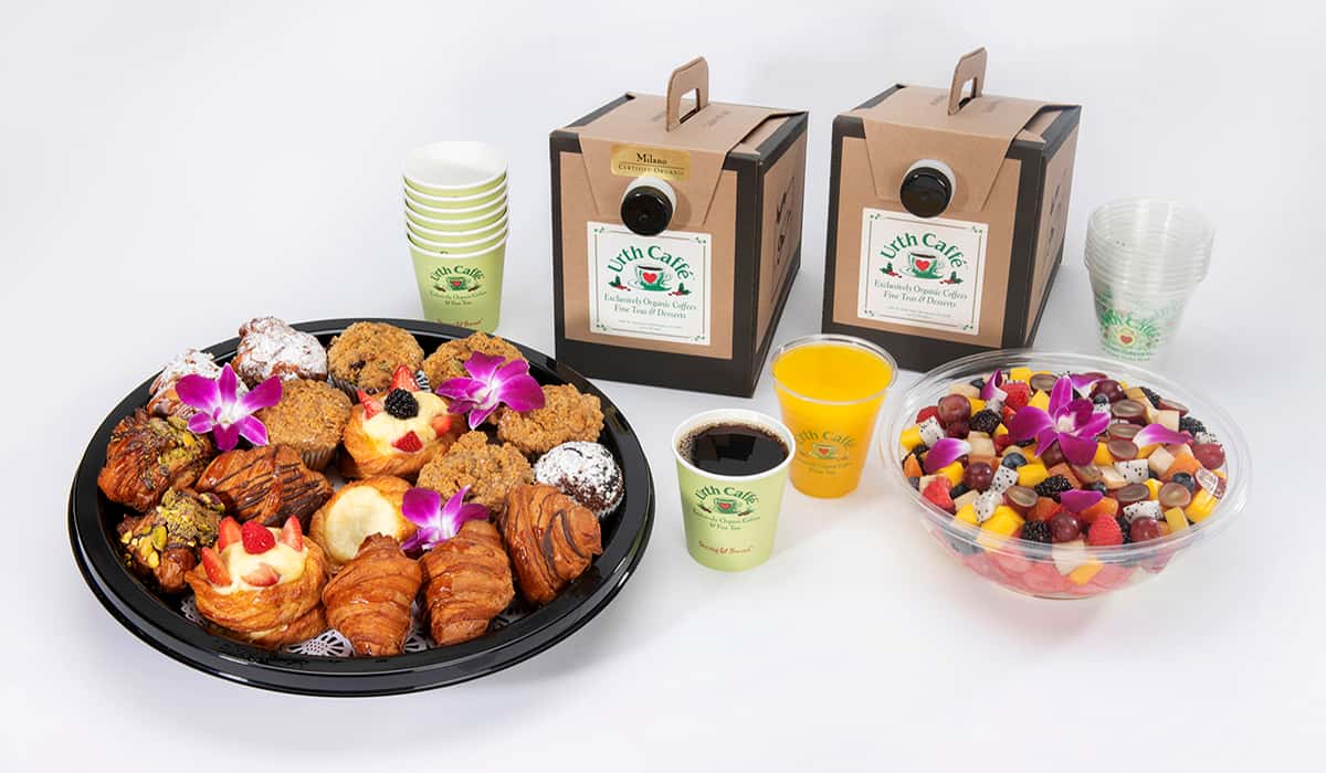Urth Catering Breakfast option showing a round platter of assorted pastries, fruit bowl and two rectangular beverage dispensers, one with a cup of coffee in front and one with a clear plastic cup of orange juice in front.