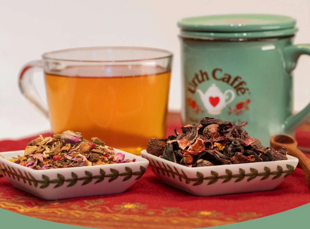 Glass of tea and green mug with Urth tea pot logo. In foreground is two little square decorative dishes with piles of different loose leaf teas