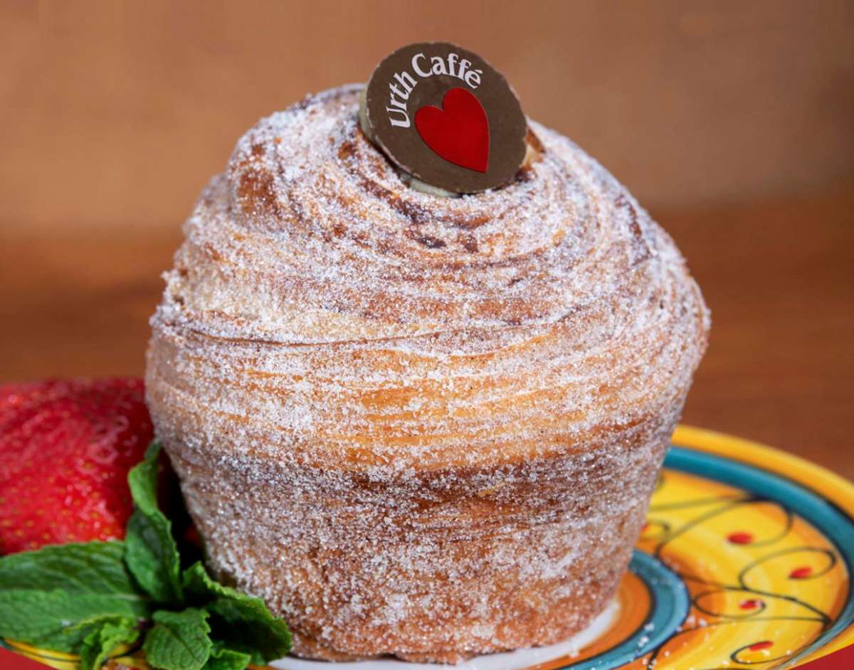 Churro Delight is a muffin shaped twirl of flaky croissant pastry with a heavy dusting of fine cinnamon and sugar. Topped with a dark chocolate coin with Urth Caffé white type arched over a red heart. It sits on a bright yellow painted ceramic plate garnished with green mint. Background is a medium wood brown.