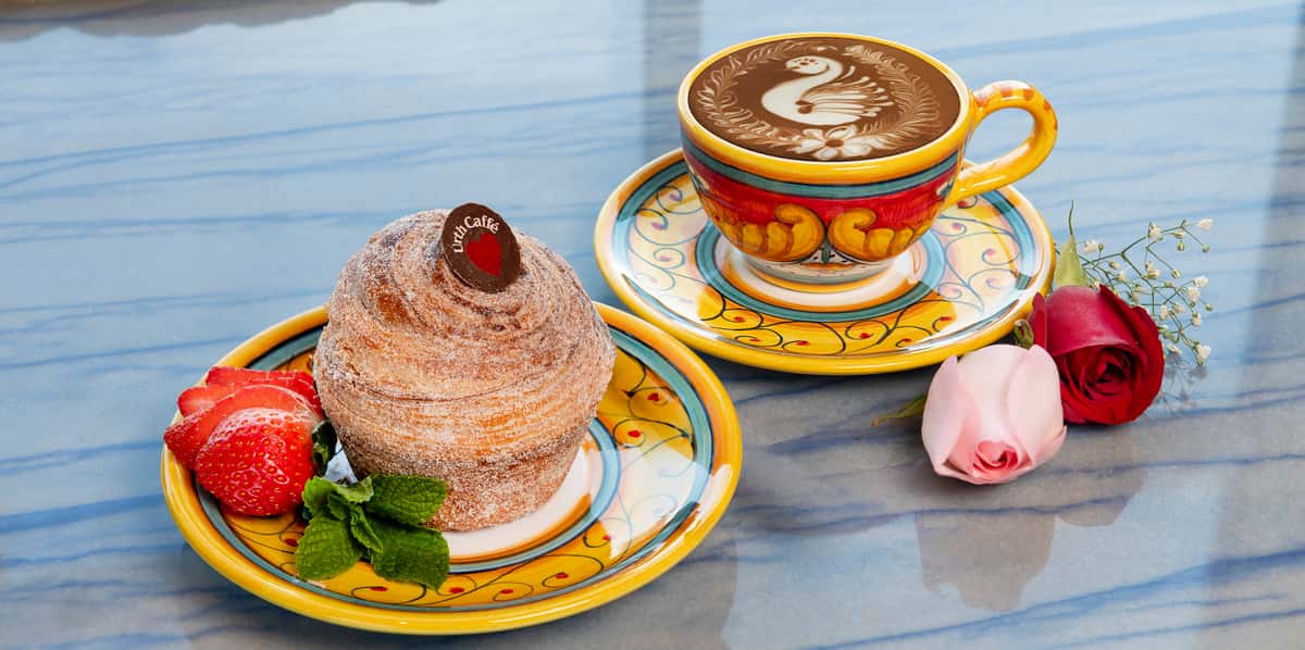 Photo of Urth's new Churro Delight a muffin shaped twisted pastry dusted in cinnamon and sugar and topped with a coin of dark chocolate. The coin has Urth Caffé printed in white and arching over a dark red heart. The churro sits on a colorful hand painted yellow saucer with a garnish of green mint and slices of red strawberry tucked against the left side of the churro. In the background, to the upper right is a matching hand painted cup and saucer. The cup has a beautiful hot cocoa latte design of a swan encircled by two fern leaves. The counter top is a blue veined polished marble.