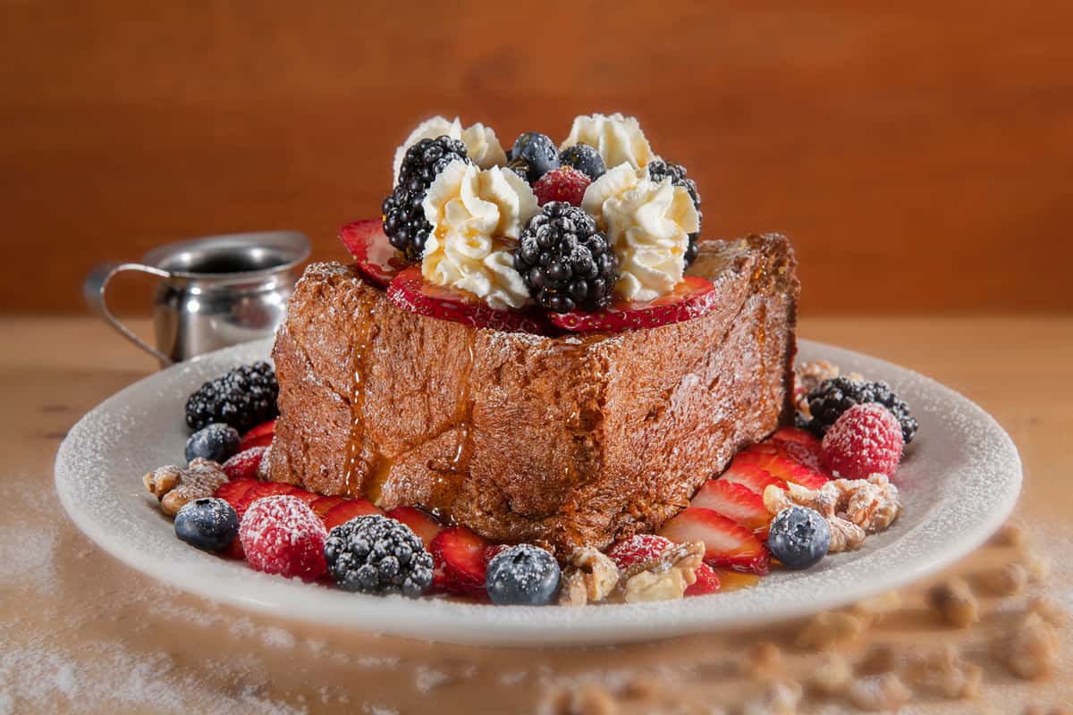 Urth's new super thick house-made brioche French Toast, baked and lightly caramelized with honey butter served with fresh whipped cream, berries and sliced strawberries, walnuts and pure maple syrup on a plate.
