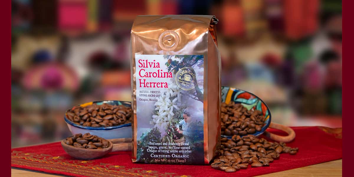 Photo of a labeled copper colored 12oz. bag of Urth Caffé's Silvia Carolina Herrera Natural-Process, Geisha Micro Lot from Chiapas, Mexico. The bag is sitting on a red cloth mat with coffee beans spilling out of a colorful ceramic bowl on the left side of the bag. Two smaller ceramic bowls full of beans sit to the left and back of the bag.