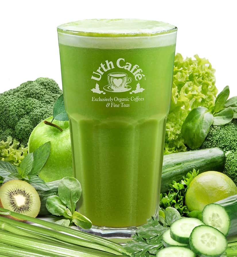 Glass of green juice with a variety of green fruits and vegetables in background