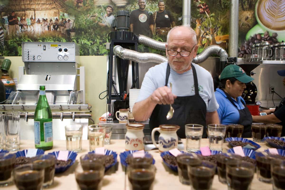 Man in an apron holds spoon over coffee cup with sample