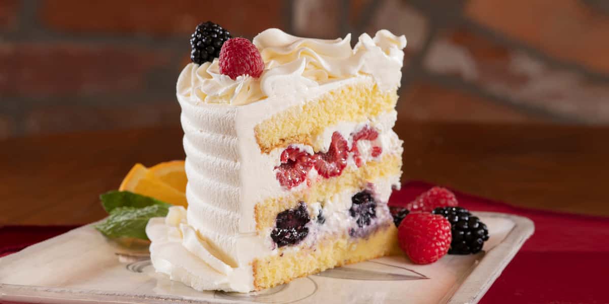 Beautiful close up of a slice of Urth's Berries and Cream cake. The white icing outside is visible to the left, raspberries and blackberries and wavy white icing is decorating the top as the expose interior is angled away from the front to the right. The interior has the three layers of light yellow sponge cake separated by two thick layers of icing, the upper one packed with sliced whole raspberries and the lower one showing sliced whole blackberries. Cake is sitting on a square antique white plate and garnished with a couple more berries. Some mint and orange slices are peaking out behind the cake on the left while the background brown wood color.