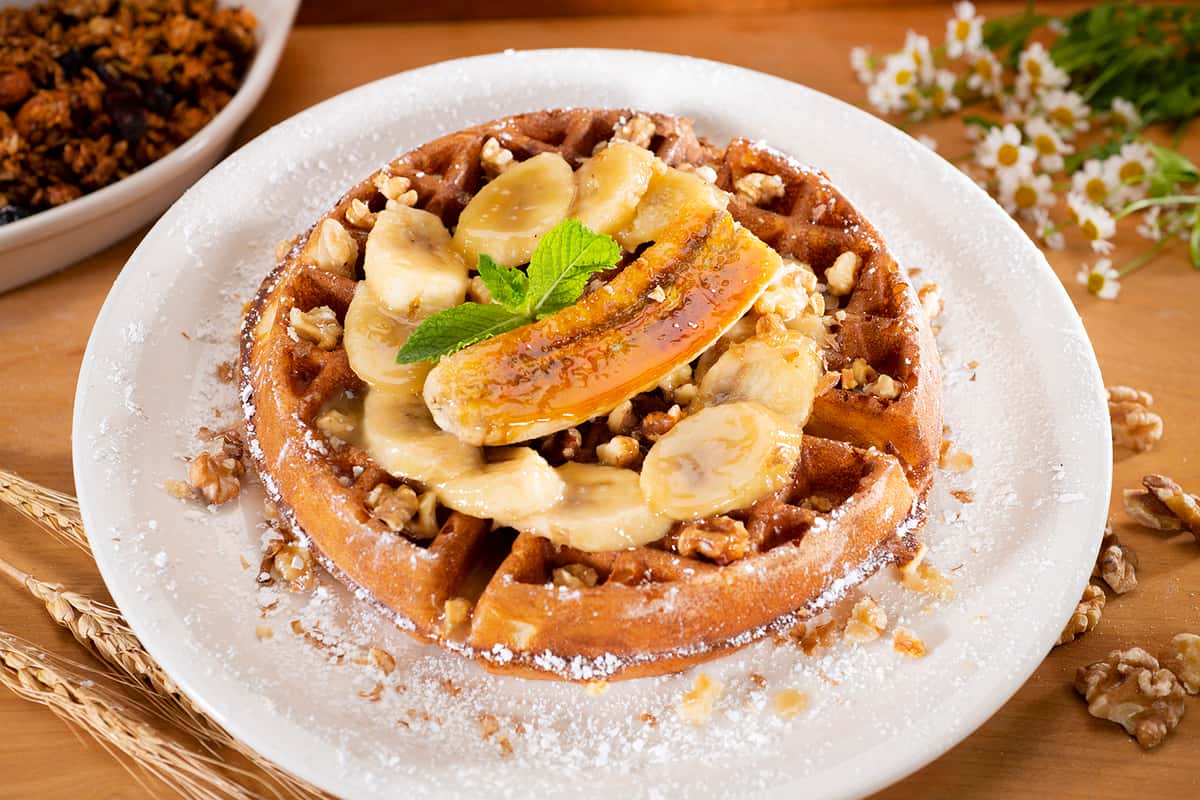White plate with a Banana Walnut Waffle topped with a caramelized banana half on top of banana slices, chopped walnuts and lightly sprinkled with powdered sugar.