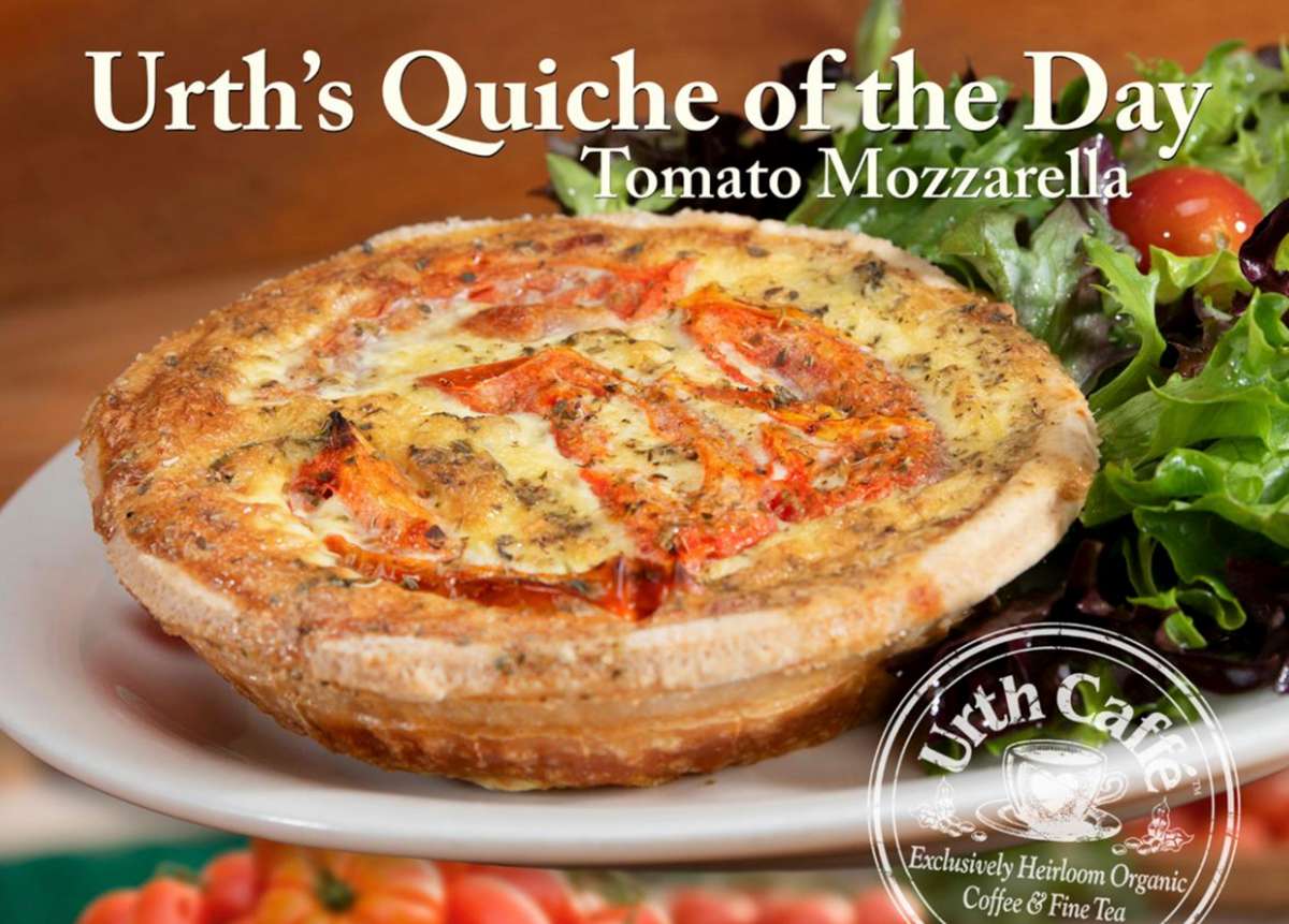 Urth's Quiche of the Day, Tomato Mozzarella. Quiche with golden crust and toasted tomatoes and mozzarella as a filling. Sitting on a white plate with a mixed greens salad behind it.
