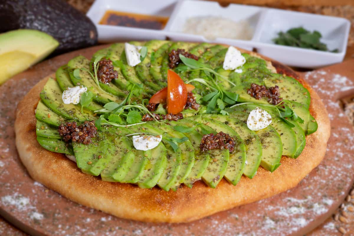 The Urth Avocado pizza with hummus, house-made tomato sauce covered with the finest fresh sliced avocado garnished with almond cheese, micro cilantro, and quinoa.