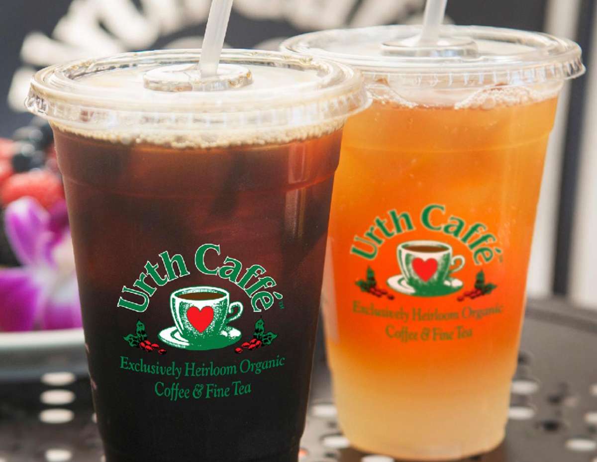 Two clear To Go Cups with color Urth Caffé logos, the one in front is a dark iced coffee color and the slightly behind it to the right is filled with orange iced tea and has lemonade at the bottom.