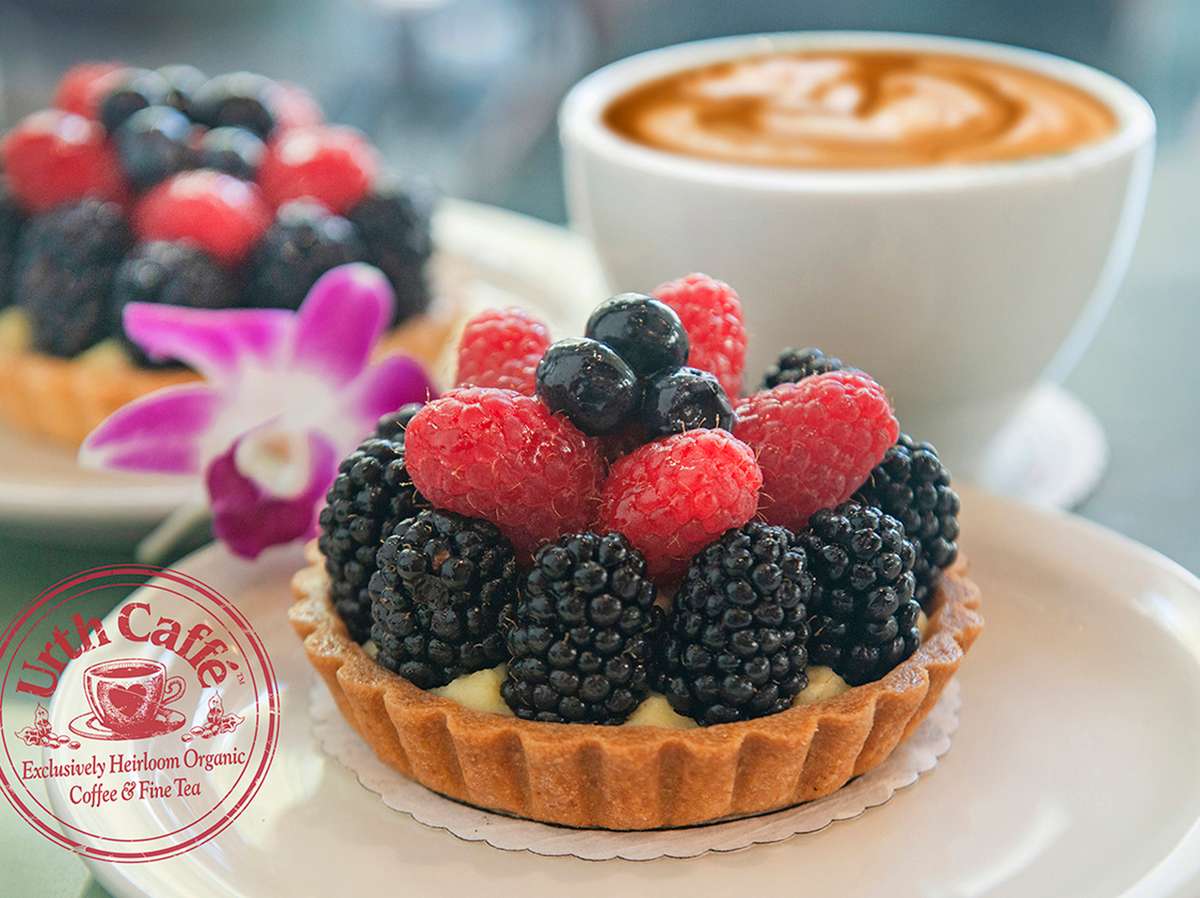 Urth Caffé Fruit tart generously topped with a row of blackberries, Raspberries and some blue berries at the top. Urth circular stamp logo is in lower left corn in dark red. Being the tart on the left is a purple orchid and another tart. the background is a white coffee cup with a latté design.