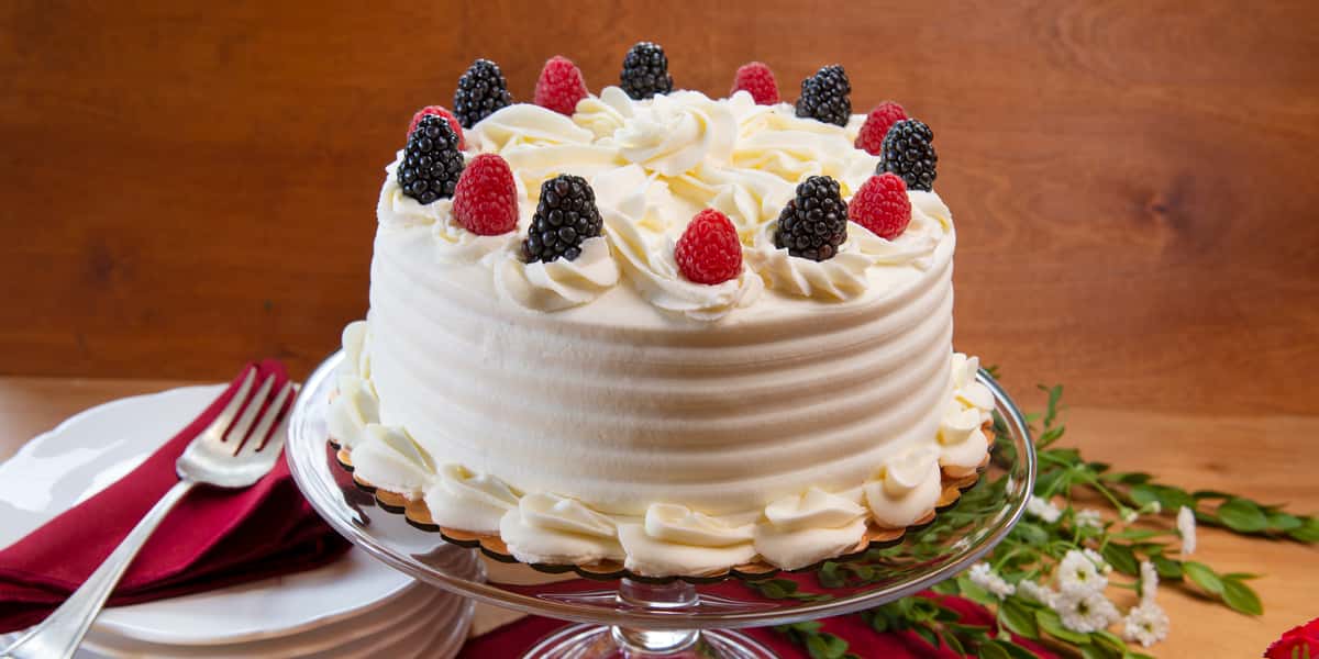 Urth Berries and Cream whole cake with all white icing and raspberries and blackberries decorating the top. Cake is sitting on a raised glass pedestal platter. Background brown wood.