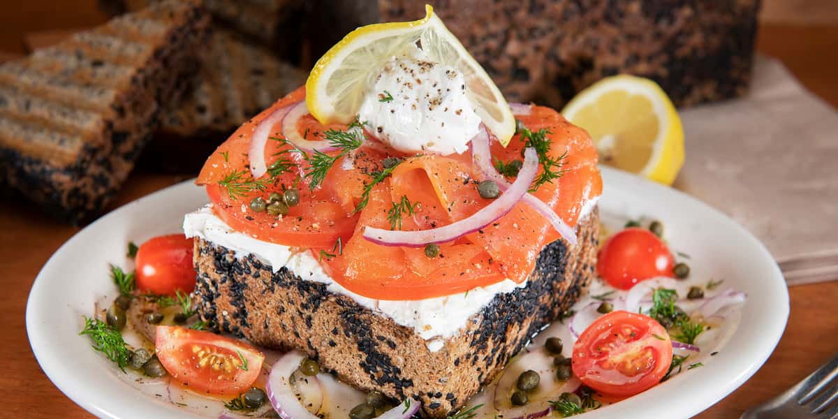white plate garnished with sliced tomatoes and capers with a thick slice of buckwheat toast topped with Lox, cream cheese, capers, thinly sliced red onion and a dollop of lemon dill mascarpone.