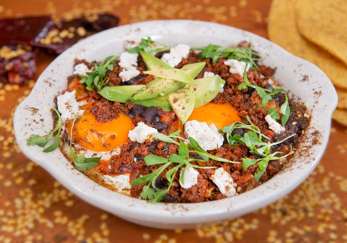 Huevos Rancheros in white bowl with eggs, beans and soy chorizo topped with almond cheese, ripe avocado and micro cilantro.