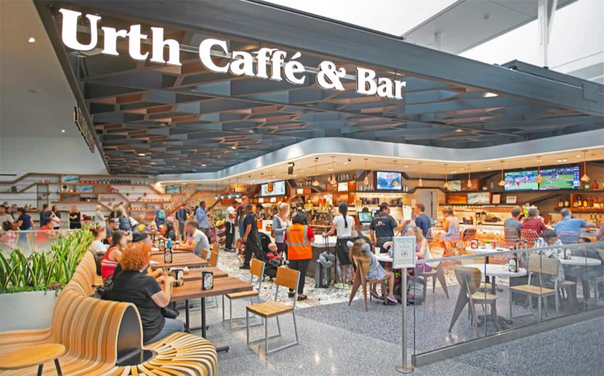 Beautiful shot of the busy Urth Caffé & Bar location at LAX. The name is in large white letters on the front edge of a grey striking angular ceiling over a undulating aluminum soffit with TV screens hanging down. Guest are sitting at the bar and at an array of wood tables and chairs while others are standing in line waiting to order.