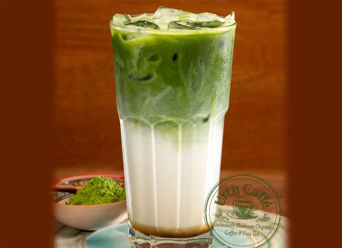 Urth Caffé's Matcha Latté over ice in a tall glass. The top is matcha green with ice and midway fades to white in the bottom half of the glass. To the left of the glass is a small bowl of powdered matcha. To the bottom right is the circular Urth Caffé stamp logo in matcha green.