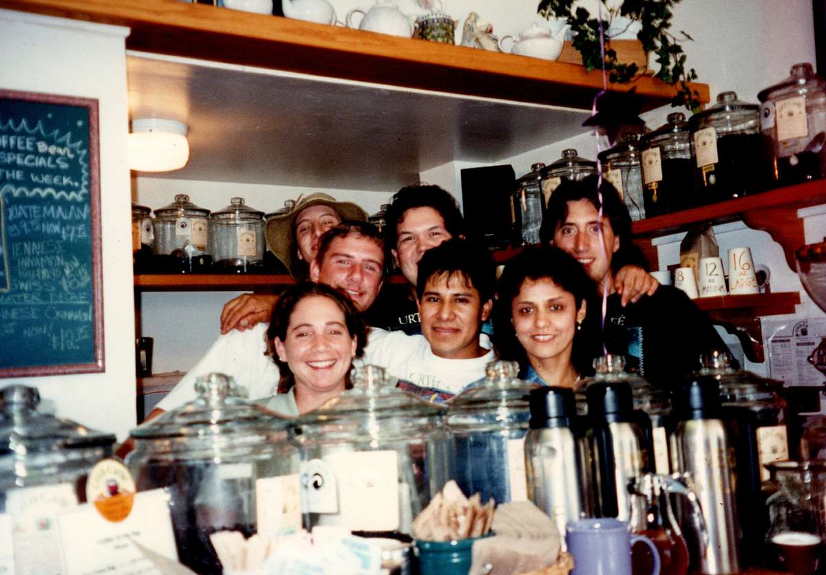 Group of 7 people behind coffee house counter with glass canisters on top of it.