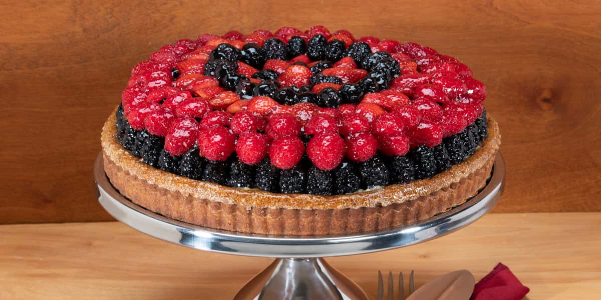 Beautiful whole 12 inch Fruit Tart showing the ridged crust topped with carefully placed layers of rows of alternating glazed black berries and red raspberries. Shown against a brown wood colored background on a raised silver pedestal dish with a fork and napkin barely visible on the table top below.