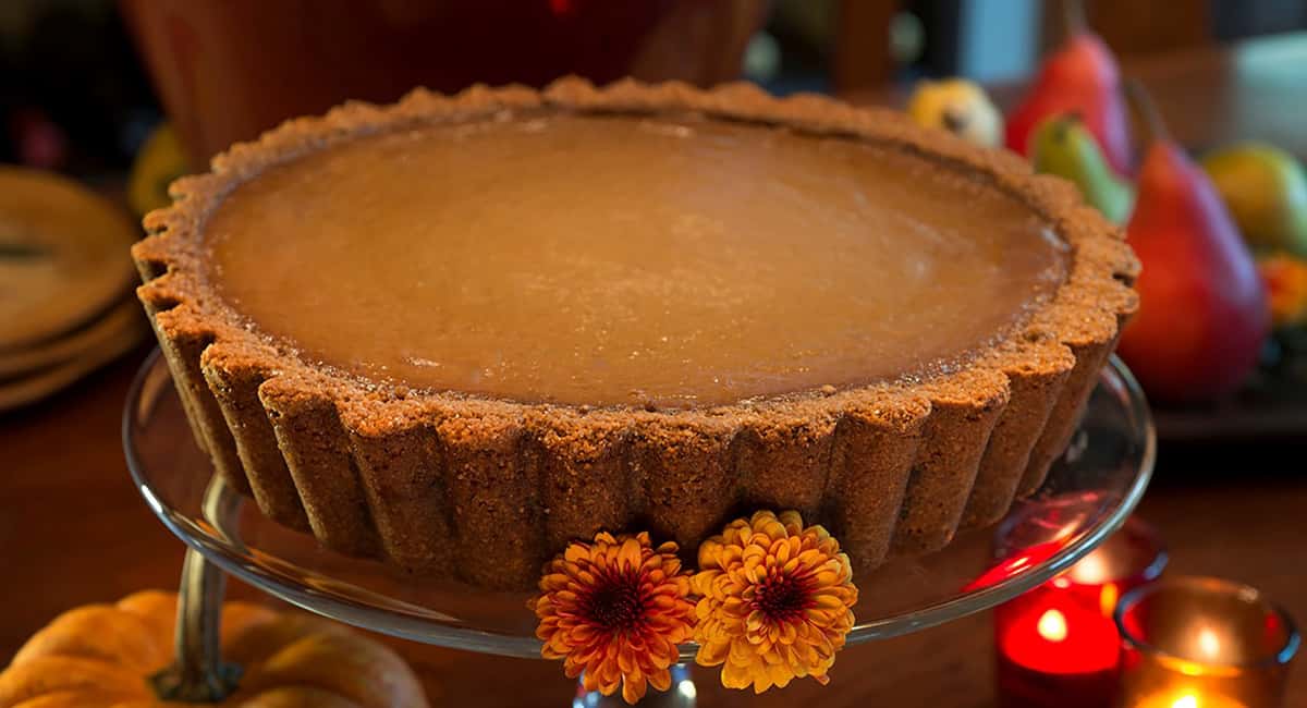 Urth’s famous thick golden brown whole Pumpkin Pie with its thick scalloped outer crust on a raised glass dish garnished with yellow and orange flowers.