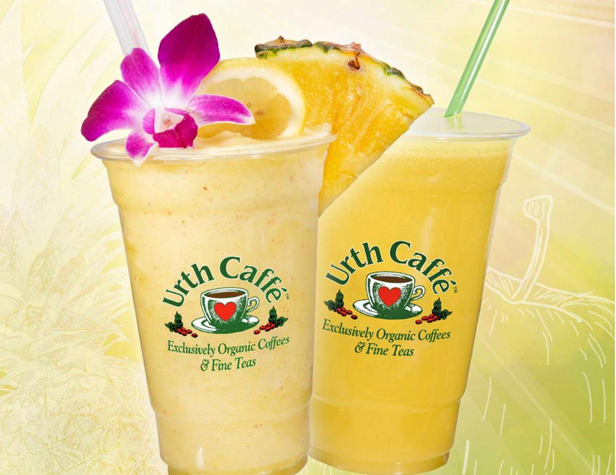 Two tilted Urth clear Urth To Go cups with Urth logos. One on the right is full of a light yellow smoothie and garnished on top with a magenta orchid and slice of pineapple. The Juice to the right is a darker yellow and has a green straw.