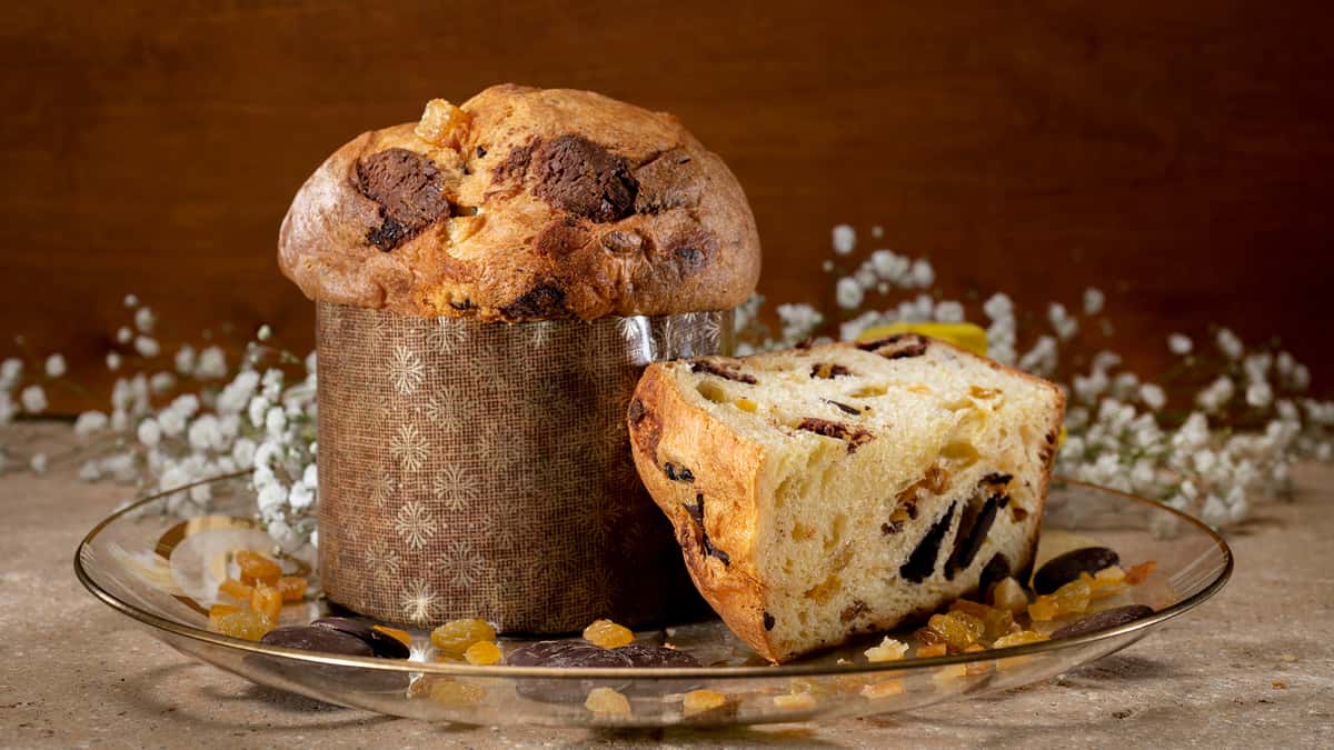 Photo of  full loaf of the Urth Valentine Panettone with chunks of Ecuadorian chocolate. A quarter slice leaned up horizontally against it to the right showing the interior contents of the bread. Chocolate and candied orange pieces are scattered around the base along with tiny white straw flowers.
