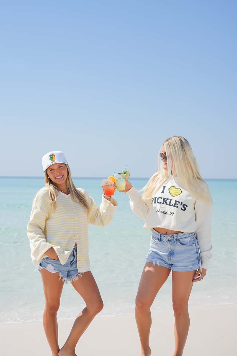 Two women toasting colorful cocktails on the beach, celebrating a fun and relaxed coastal dining experience.
