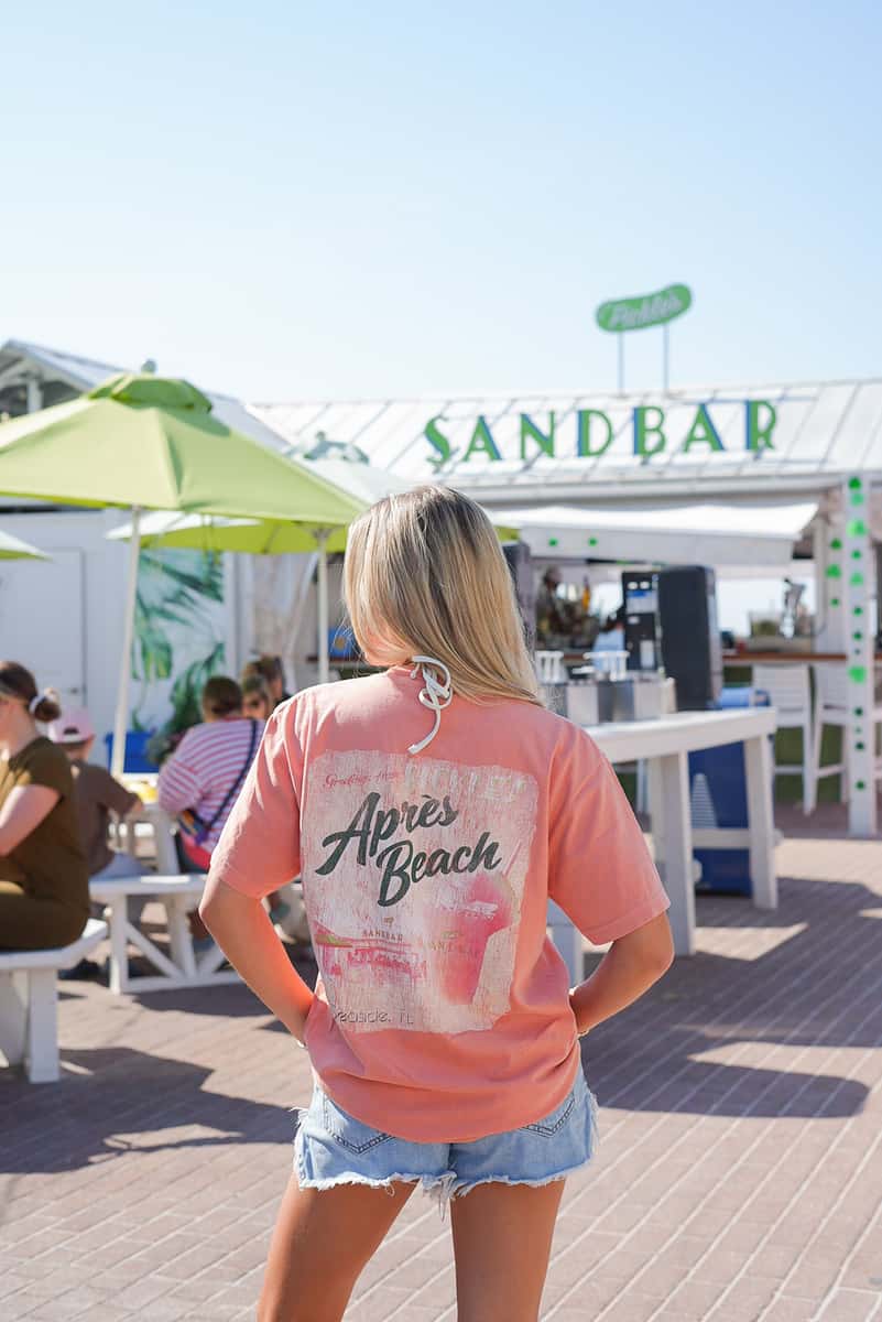 Guest wearing a Sandbar "Après Beach" T-shirt on the outdoor patio in front of the waterfront restaurant, showcasing the restaurant's coastal lifestyle and branded merchandise.