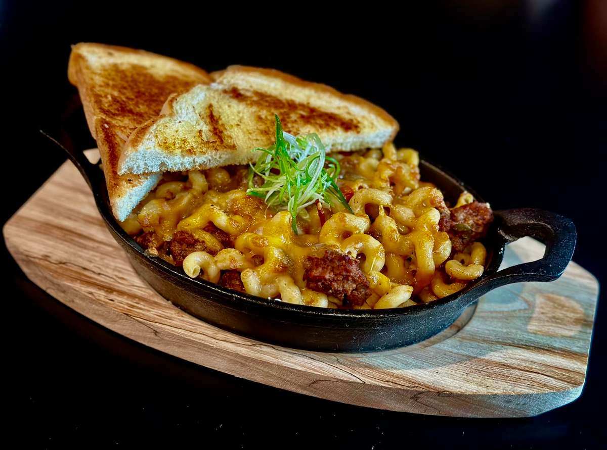 A serving of macaroni and cheese with ground beef and melted cheddar cheese, served in a cast-iron skillet, accompanied by two slices of toasted bread. The dish is garnished with green onions.