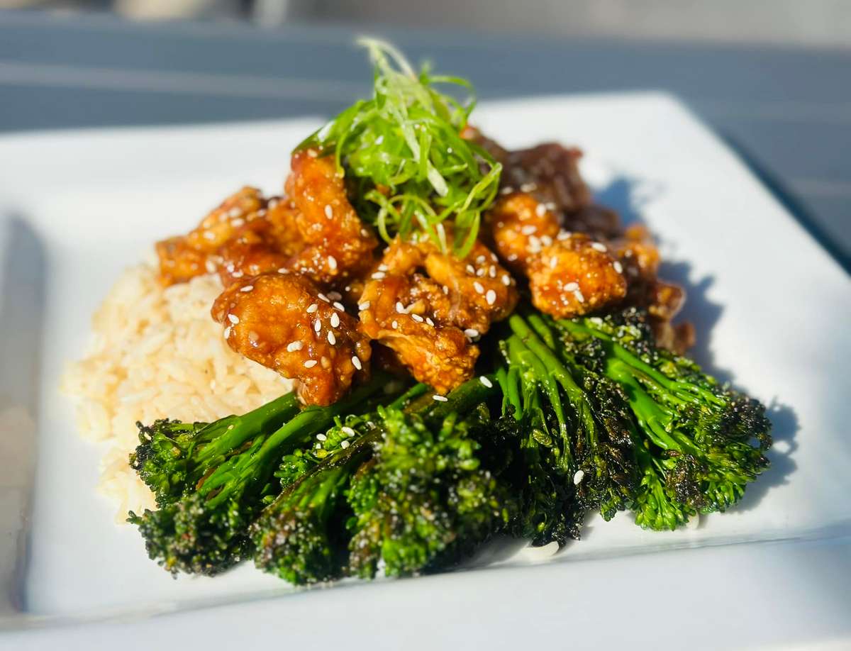 A plate with crispy chicken pieces coated in a sweet sauce, served with rice, sautéed broccolini, and garnished with sesame seeds and green onions.