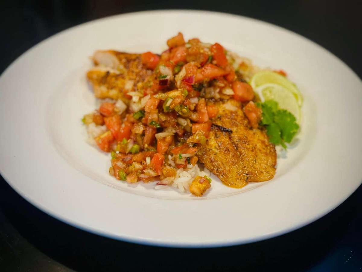 A plate of grilled fish topped with fresh tomato salsa, served over rice with lime wedges and cilantro garnish.