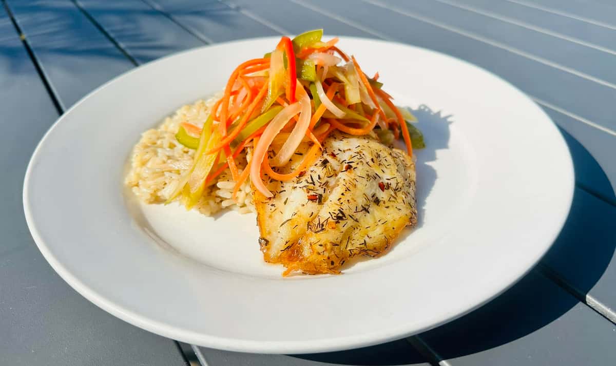 A plate with seasoned fish fillet, topped with a tangy mixture of pickled vegetables, served with rice. The dish is set on a light-colored table.