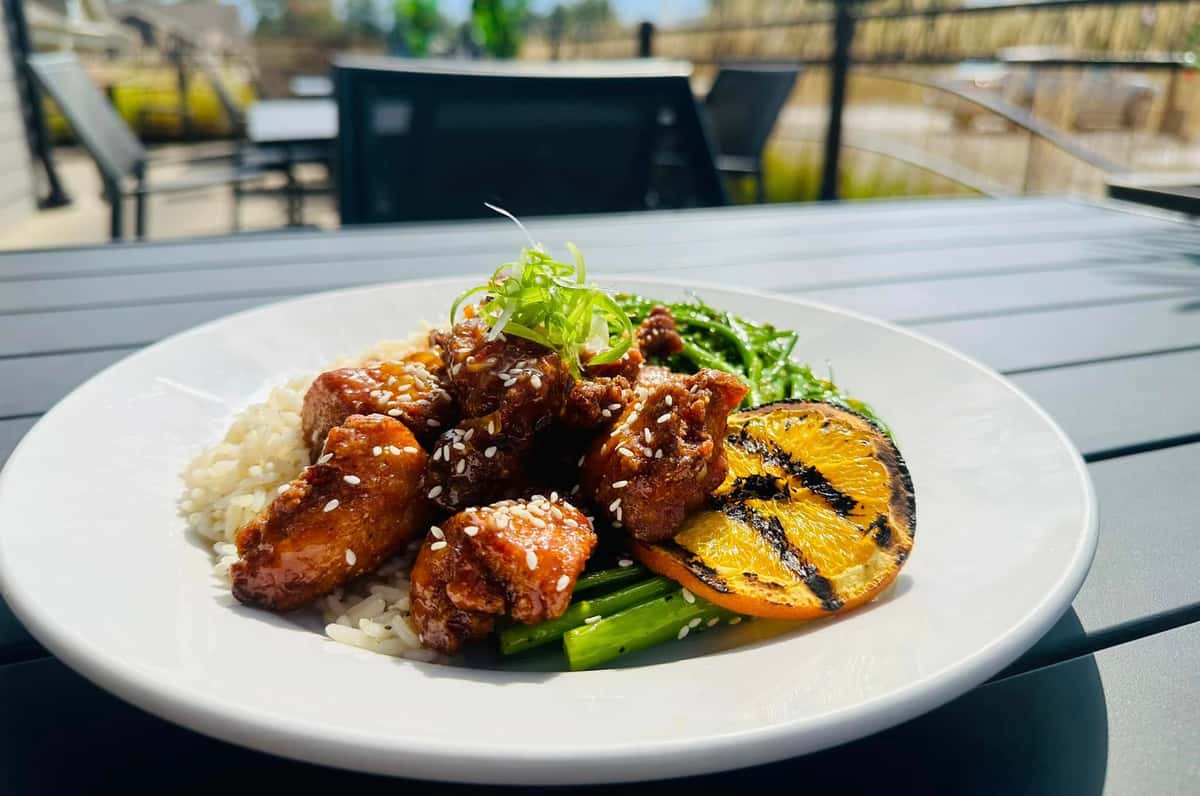 A dish featuring sesame chicken served with rice, grilled vegetables, and a charred orange slice, set on a table with an outdoor view.