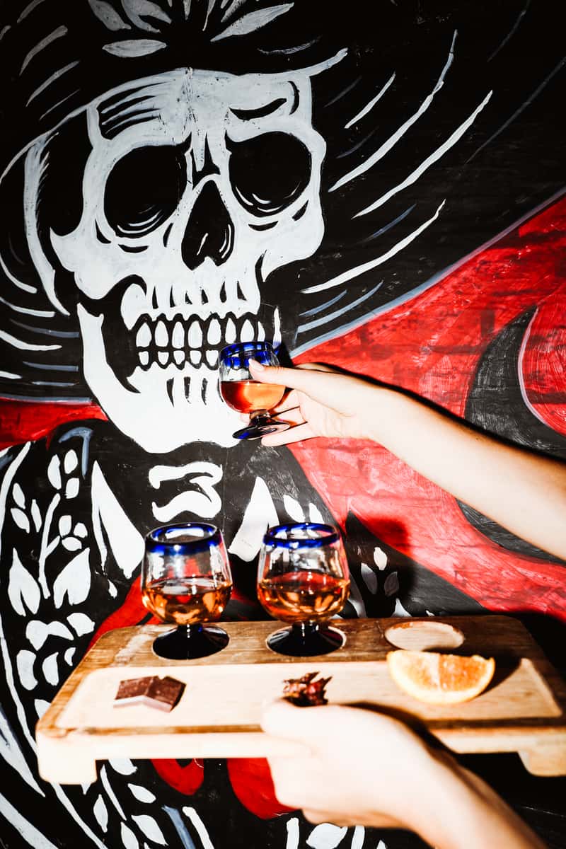 Waitress holding a liquor flight, hilariously offering a sugar skull mural a sip of one of the cocktails