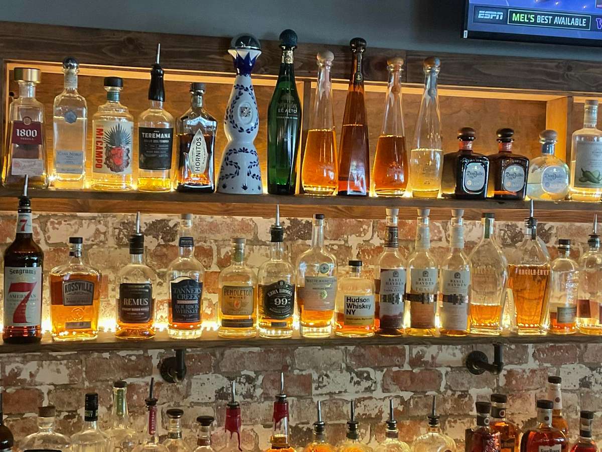 Backlit bar shelves displaying premium tequila, whiskey, and bourbon bottles against exposed brick, creating a warm upscale cocktail bar ambiance.