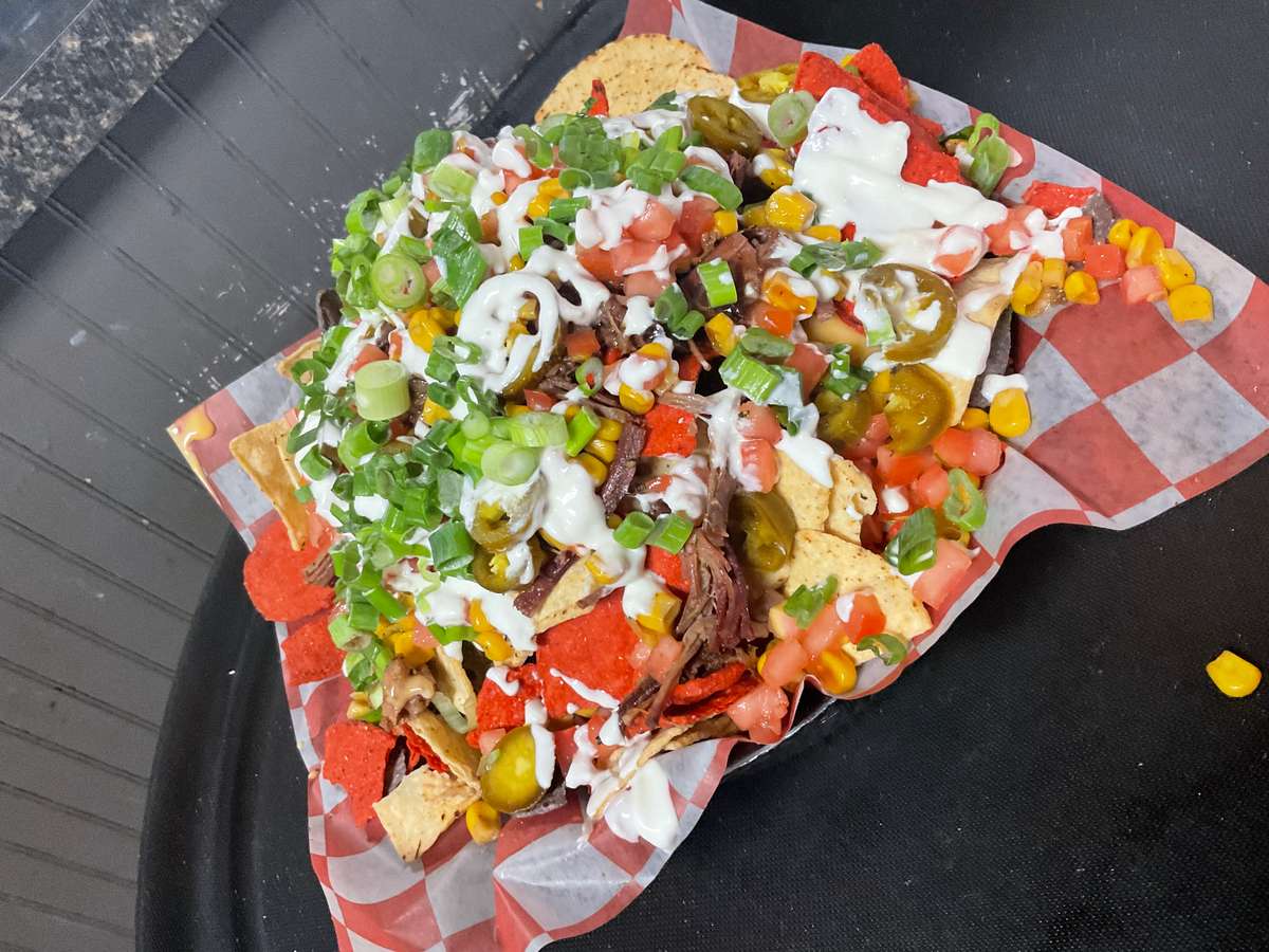 Loaded nachos topped with shredded beef, jalapeños, diced tomatoes, corn, green onions, Doritos chips, and creamy white sauce served on a checkered paper liner.