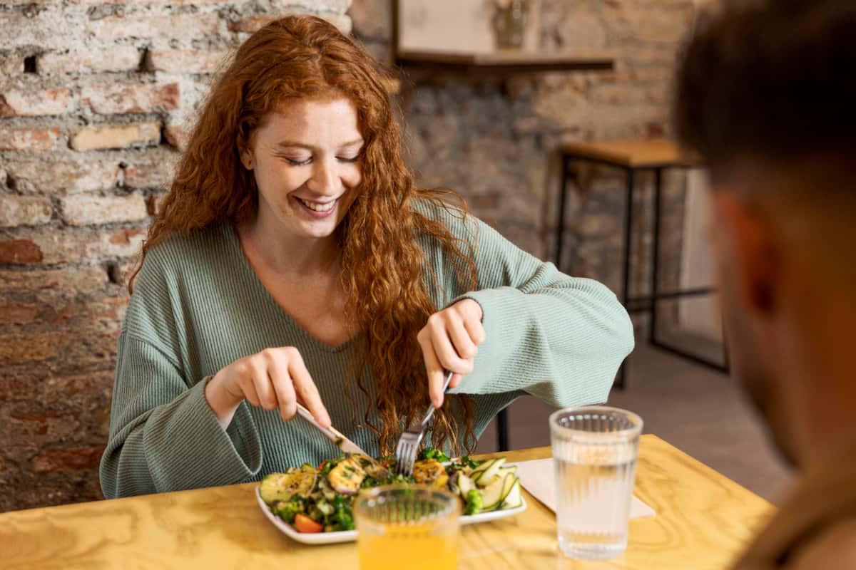 A smiling woman with curly red hair enjoys her meal at a restaurant, using a fork and knife to eat a salad, seated at a wooden table