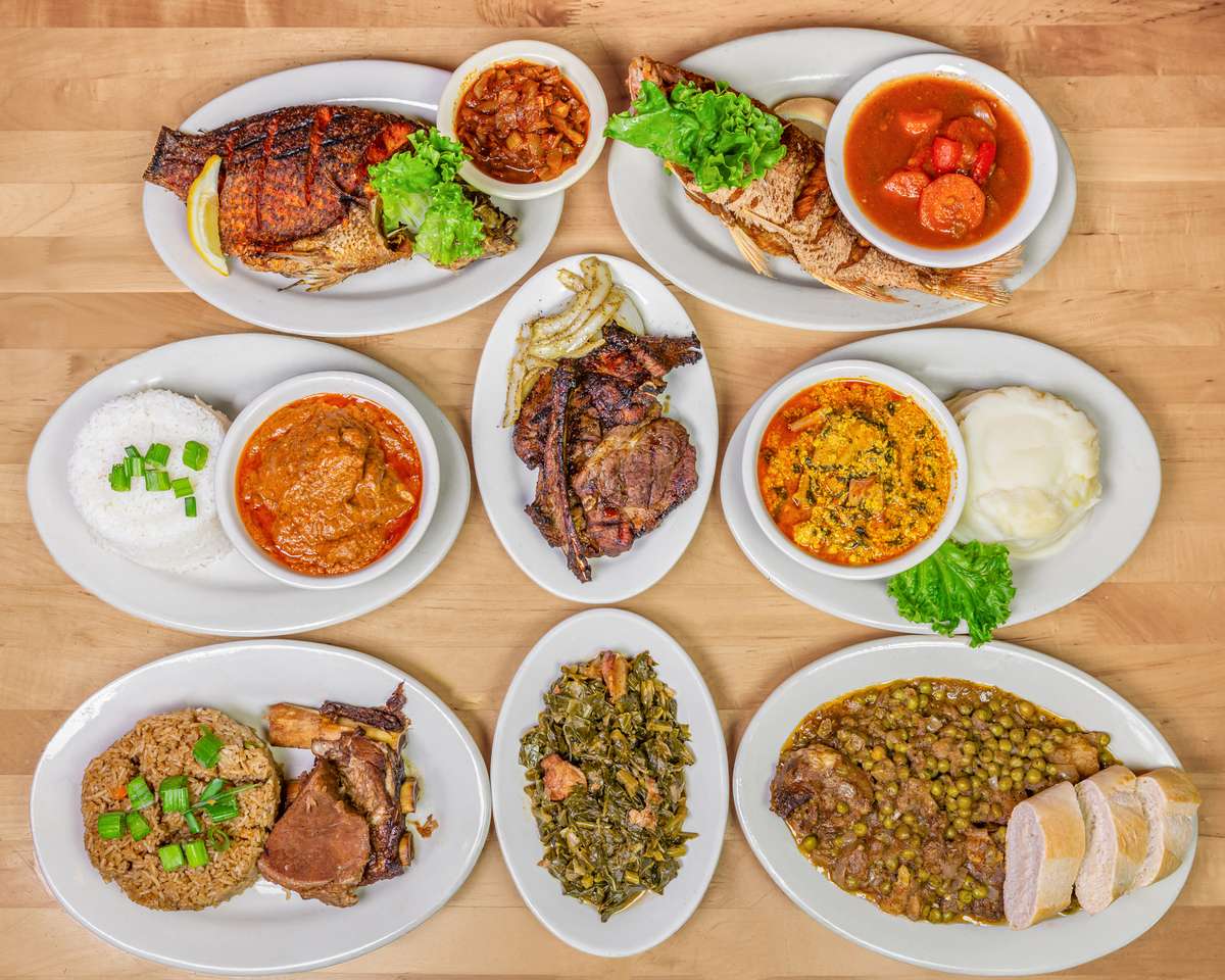Colorful spread of authentic West African dishes featuring grilled whole tilapia with fresh lettuce and lemon garnish, spicy tomato stew, egusi soup with fufu, jollof rice with lamb, pepper soup, sautéed greens, grilled goat meat, and a hearty pea stew served with sliced bread, all artfully arranged on a wooden table.