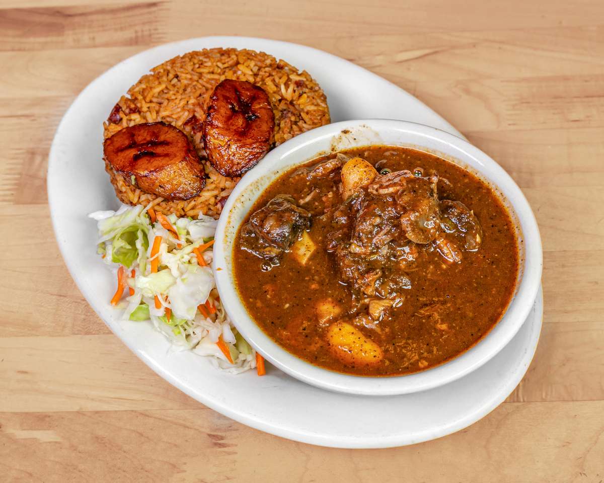 Plate of jollof rice topped with fried plantains, served with a bowl of hearty West African pepper soup loaded with assorted meats and root vegetables, accompanied by fresh cabbage slaw.