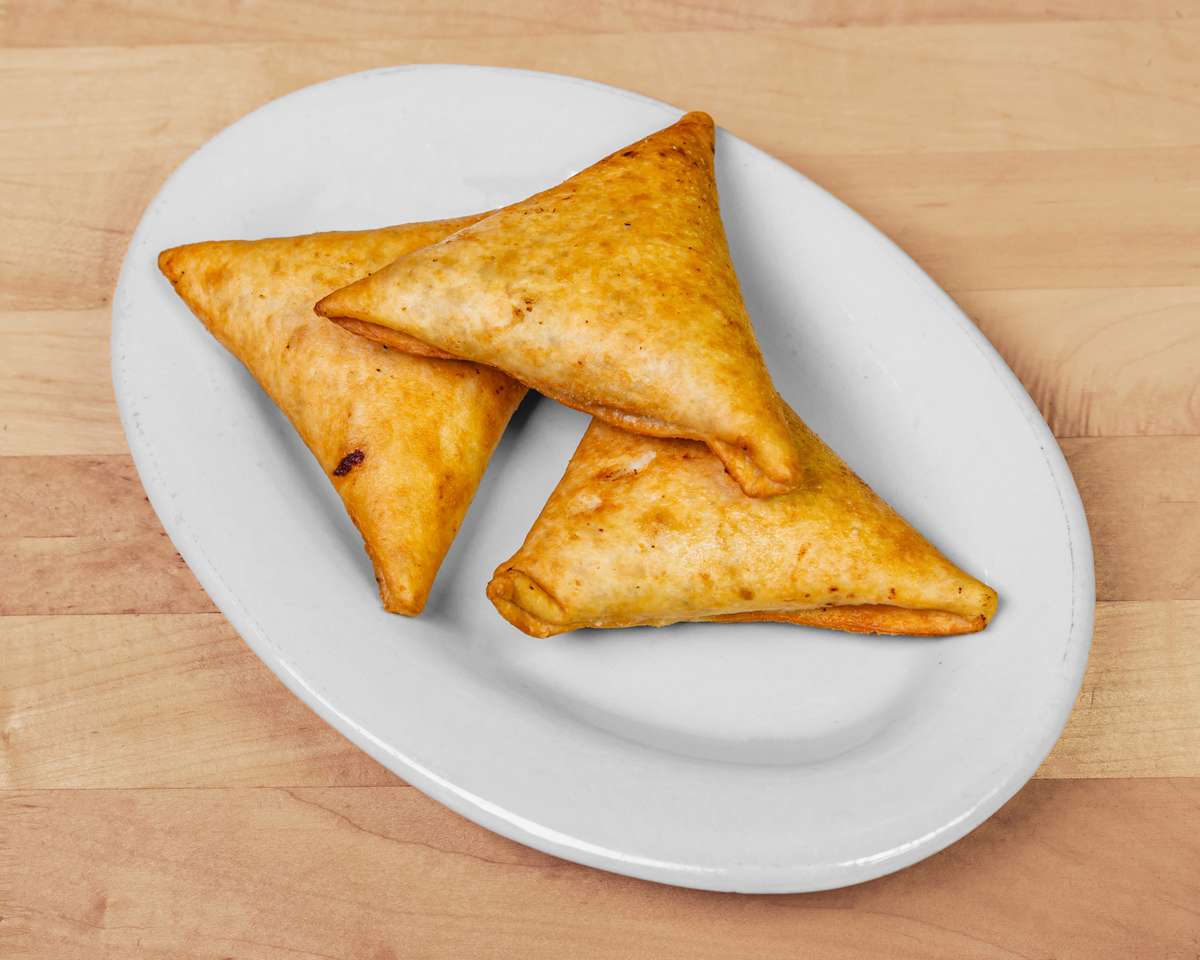 Trio of golden-brown fried samosas served on a white plate — crispy pastry pockets typically filled with spiced vegetables or meat, popular in Caribbean and South Asian cuisine.