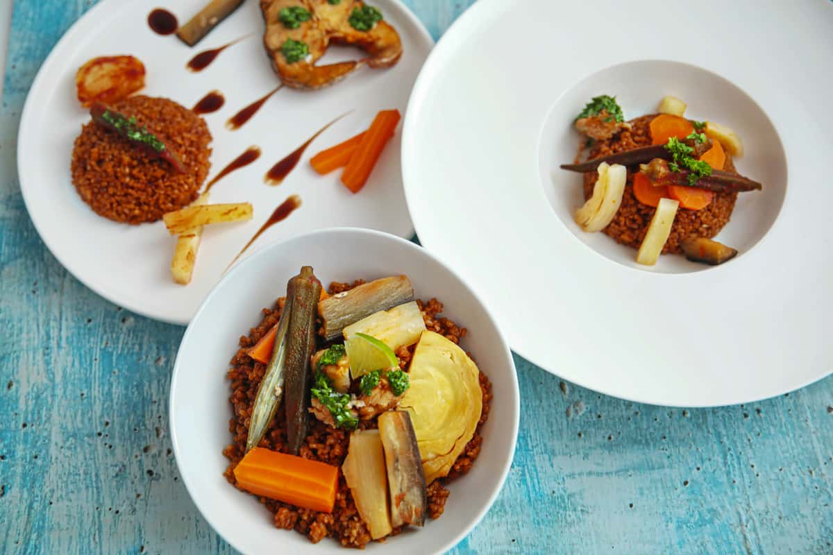 Elegant presentation of fonio grain with assorted steamed root vegetables, okra, and cabbage, served in minimalist white bowls and plates, styled on a blue rustic background.