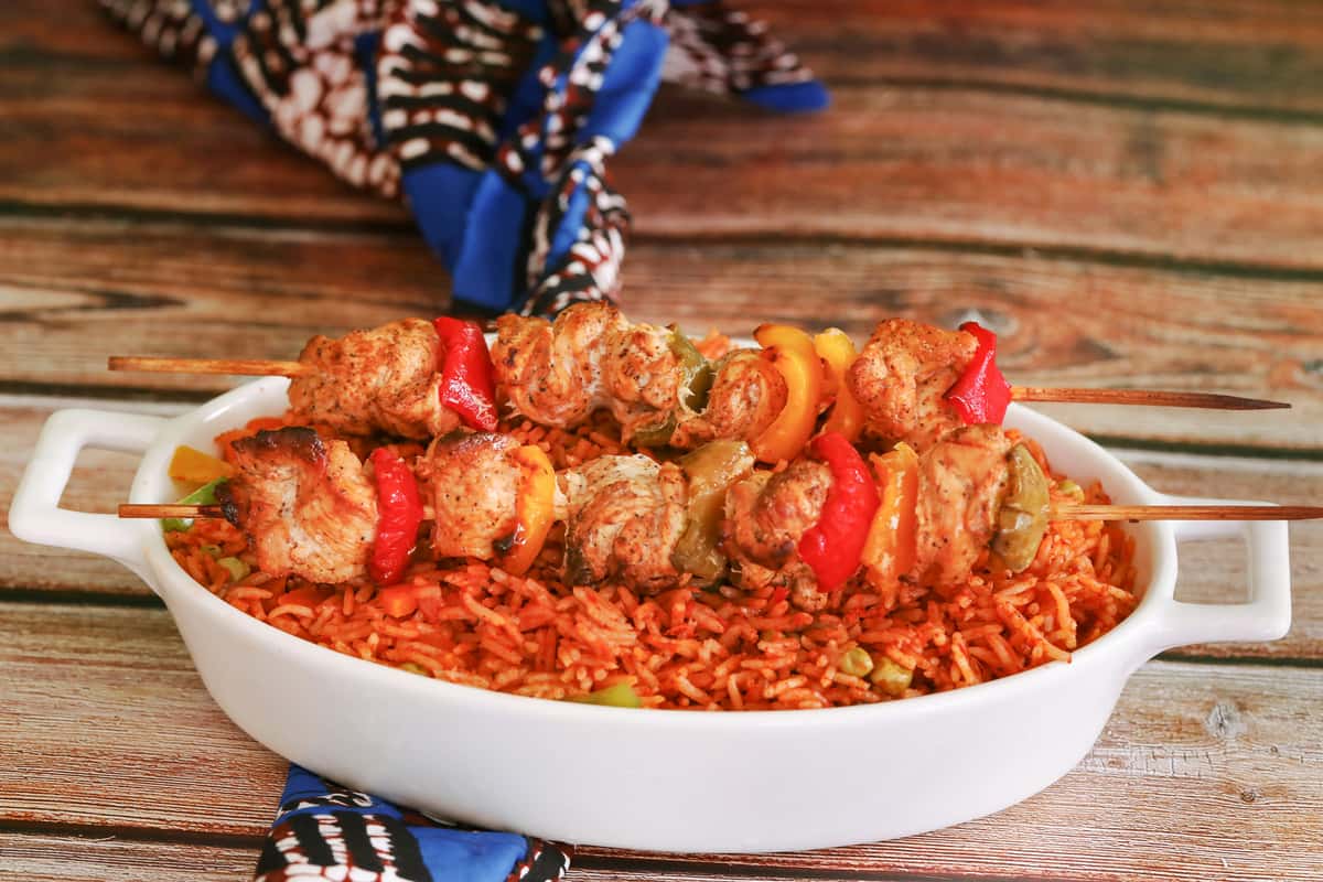 Nigerian jollof rice served in a white dish, topped with two skewers of grilled chicken and colorful bell peppers, set against a rustic wooden background with vibrant fabric.