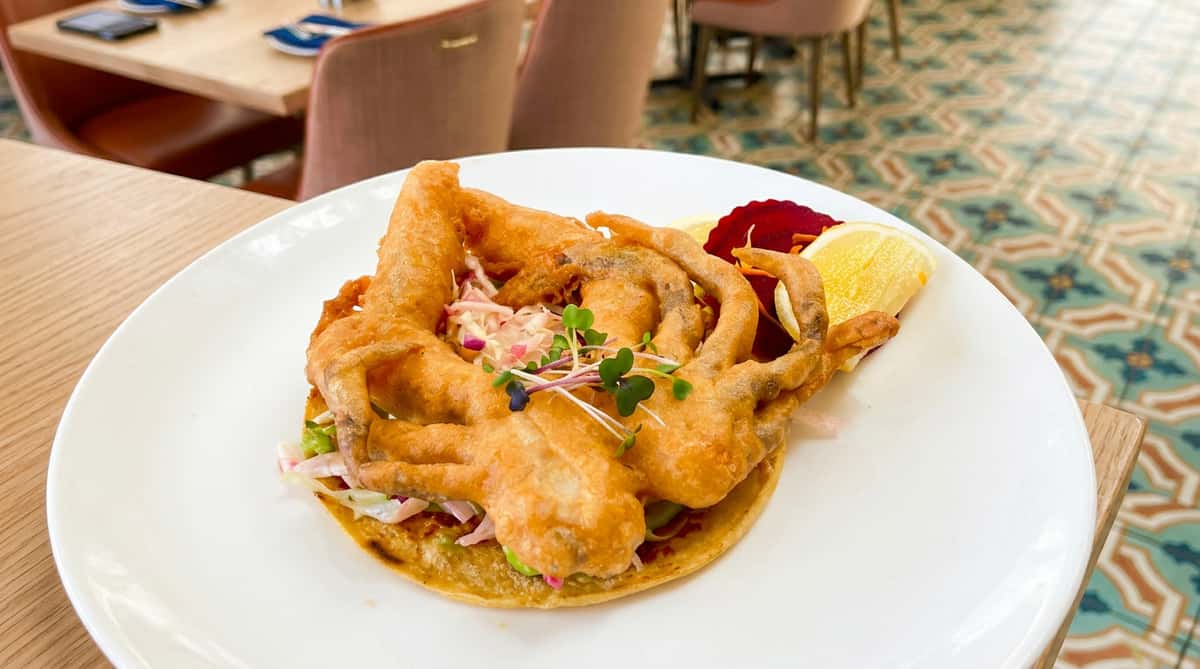 Soft-shell crab taco topped with fresh slaw and microgreens, served on a corn tortilla with pickled vegetables and a lemon wedge.