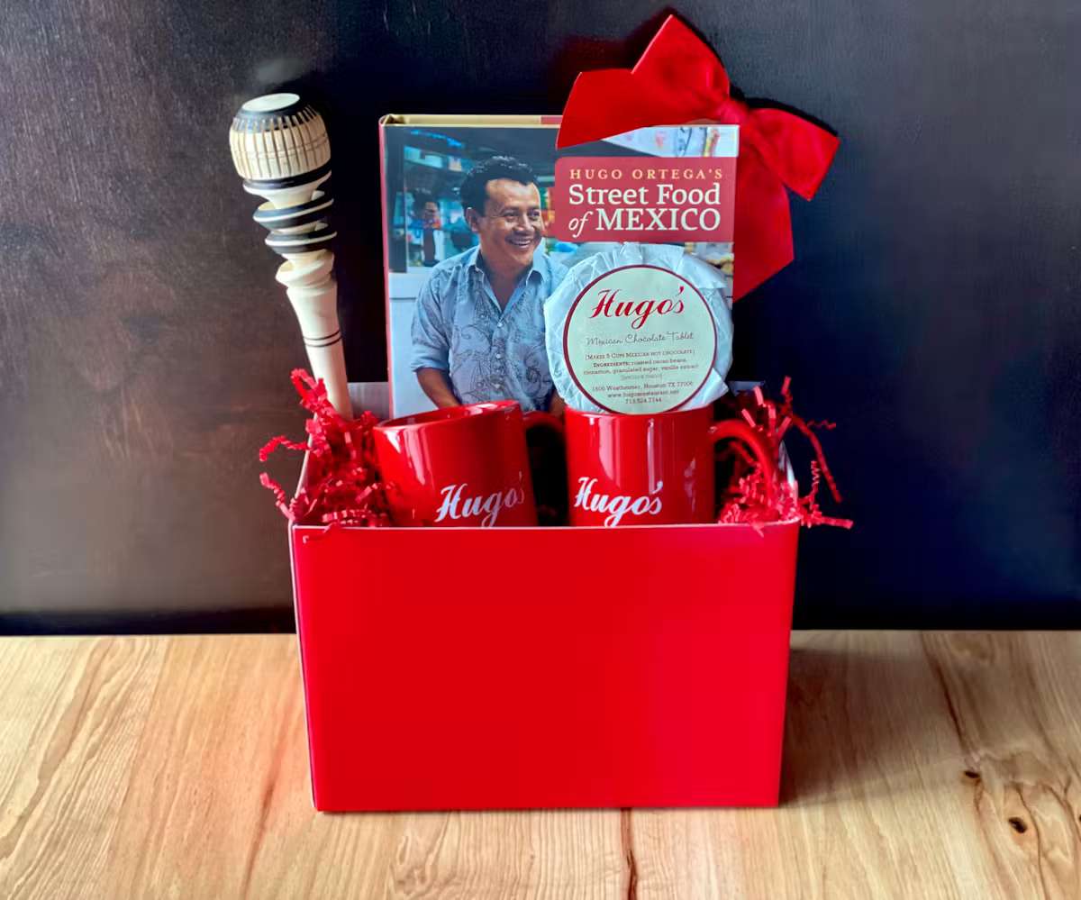 Hugo's Mexican gift basket featuring branded mugs, Mexican hot chocolate, Hugo Ortega's Street Food of Mexico cookbook, and a traditional molinillo for authentic hot chocolate.