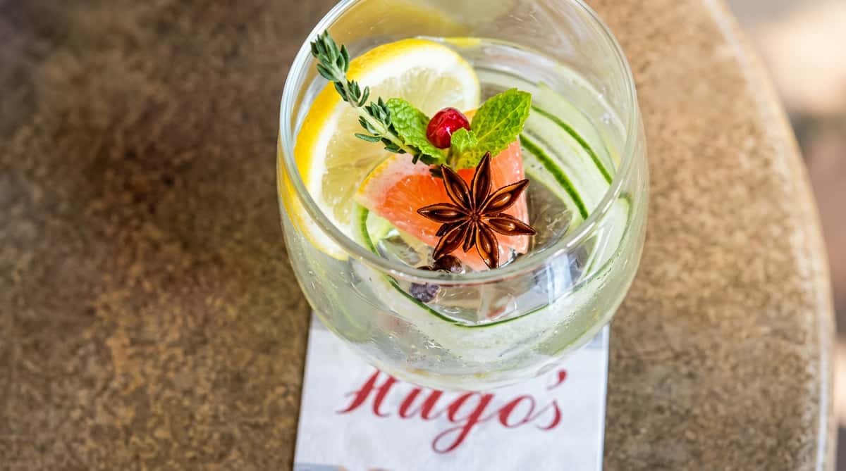Clear beverage in a large glass with lemon, cucumber slices, various herbs, a cranberry, and star anise.
