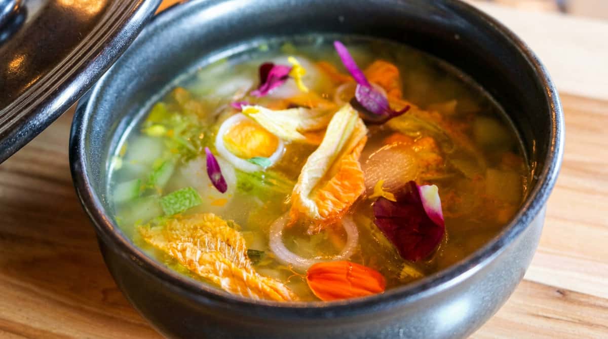 Clear soup with vegetables, egg slices, and red, yellow, and purple edible flowers in a black bowl.