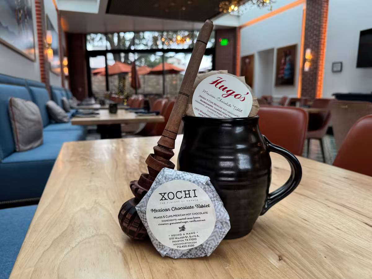 Traditional Mexican hot chocolate gift set featuring a ceramic mug, wooden molinillo whisk, and Hugo's and Xochi Mexican chocolate tablets displayed inside an elegant restaurant dining room.
