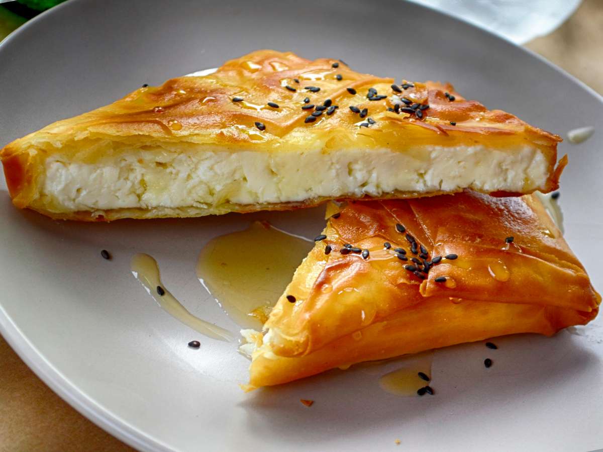 A plate of crispy phyllo pastry triangles filled with feta cheese, drizzled with honey, and sprinkled with black sesame seeds—likely a sweet-savory Greek appetizer such as "Feta Me Meli."